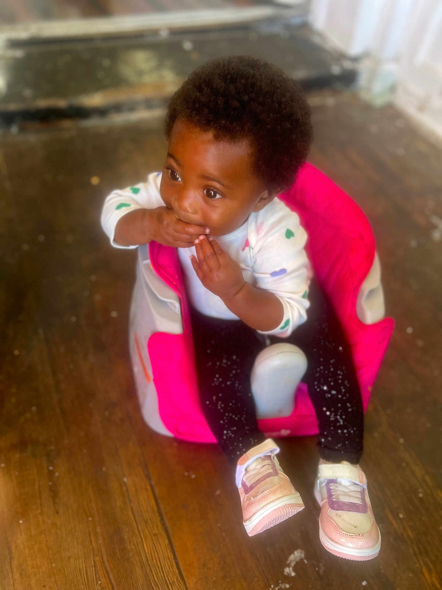 Jahmira joined the competition — help win amazing prizes! toddler, child, baby_seat, pink, white_shirt, black_pants, sneakers, wooden_floor, indoor, curly_hair, cute, sitting, hands_near_mouth, expression, young_child, footwear, floor, person, portrait, casual_clothing