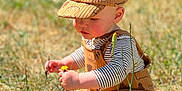 Everest is registered to the contest to win money with this photo: toddler, child, baby, grass, flower, yellow_flower, hat, cap, outdoor, nature, sunlight, barefoot, overalls, striped_shirt, playing, sitting, curious, greenery, summer, daytime