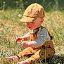 toddler, child, baby, grass, flower, yellow_flower, hat, cap, outdoor, nature, sunlight, barefoot, overalls, striped_shirt, playing, sitting, curious, greenery, summer, daytime