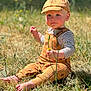 Everest is registered to the contest to win money with this photo: toddler, child, grass, outdoor, sunlight, barefoot, cap, overalls, flower, nature, striped_shirt, sitting, curious, young_child, summer, greenery, baby, portrait, daylight, casual_clothing