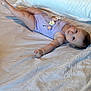 baby, infant, bed, pillow, onesie, purple_clothing, crocheted_flowers, lying_down, white_sheet, indoor, wall_outlet, curious_expression, skin, foot, hand, head, room, carpet, door_frame, soft_lighting