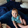 baby, infant, child, blue_onesie, cushion, bed, blanket, crib, indoor, person, skin, face, legs, arm, smile, curious, seated, furniture, room, soft_light