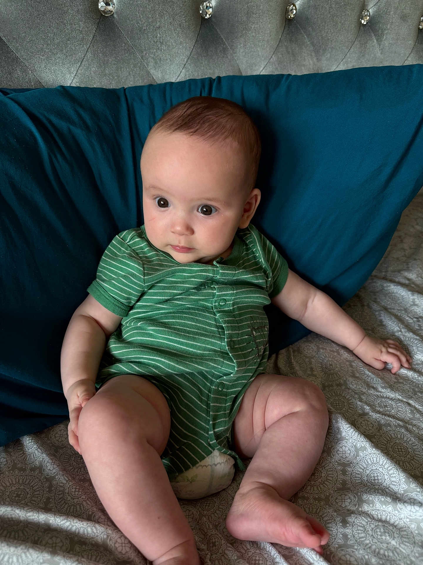 Anya is registered to the contest to win money with this photo: baby, infant, child, onesie, green_clothing, striped_clothing, bed, pillow, blue_pillow, patterned_bedspread, skin, legs, arms, face, eyes, expression, sitting, indoors, soft_fabric, cute
