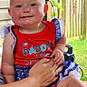 baby, child, infant, smiling, portrait, outdoor, porch, red_outfit, blue_outfit, patriotic_clothing, dress, ruffles, adult_hand, family, fence, greenery, sunlight, seated, happy, photography