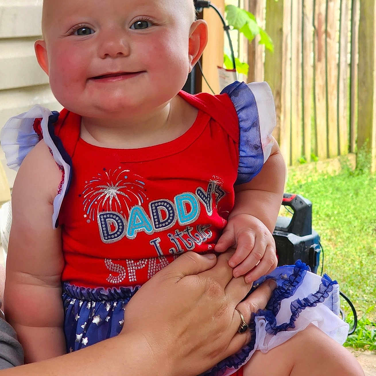 Alaina is registered to the contest to win money with this photo: baby, child, infant, smiling, portrait, outdoor, porch, red_outfit, blue_outfit, patriotic_clothing, dress, ruffles, adult_hand, family, fence, greenery, sunlight, seated, happy, photography