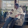 elderly_woman, dog, armchair, indoor, potted_plants, window_blinds, casual_clothing, sneakers, socks, wooden_floor, home_interior, glasses, short_hair, floral_pattern, seated, pet, cozy, natural_light, watch, bracelet
