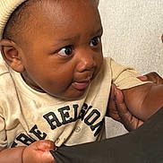 Asad is registered to the contest to win money with this photo: baby, child, infant, beanie, hat, shirt, text_on_shirt, hand, face, curious, grabbing, fabric, closeup, portrait, indoor, person, skin, expression, cute, young