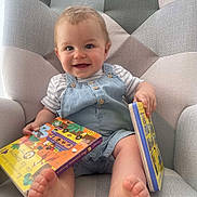 Leo a rejoint le concours — aidez-le/la à gagner de superbes lots ! baby, child, smile, book, reading, armchair, foot, clothing, denim_overall, striped_shirt, indoor, happy, cute, infant, person, face, hand, toes, furniture, playful