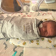 Eli is registered to the contest to win money with this photo: newborn, baby, hospital, incubator, nasal_tube, sleeping, swaddled, grey_clothing, pillow, animal_pattern, footprints, medical, infant, bed, cute, peaceful, wrapped, healthcare, premature, crib