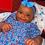 baby, child, smiling, blue_dress, headband, bow, bed, blanket, pillow, pattern, floral, happy, infant, cute, clothing, person, indoors, skin, hand, lying_down