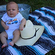 Braxton is registered to the contest to win money with this photo: baby, cowboy_hat, blanket, outdoor, grass, metal_tub, plush_toy, blue, gray, striped, sitting, infant, clothing, foot, hand, person, cute, portrait, summer, toy