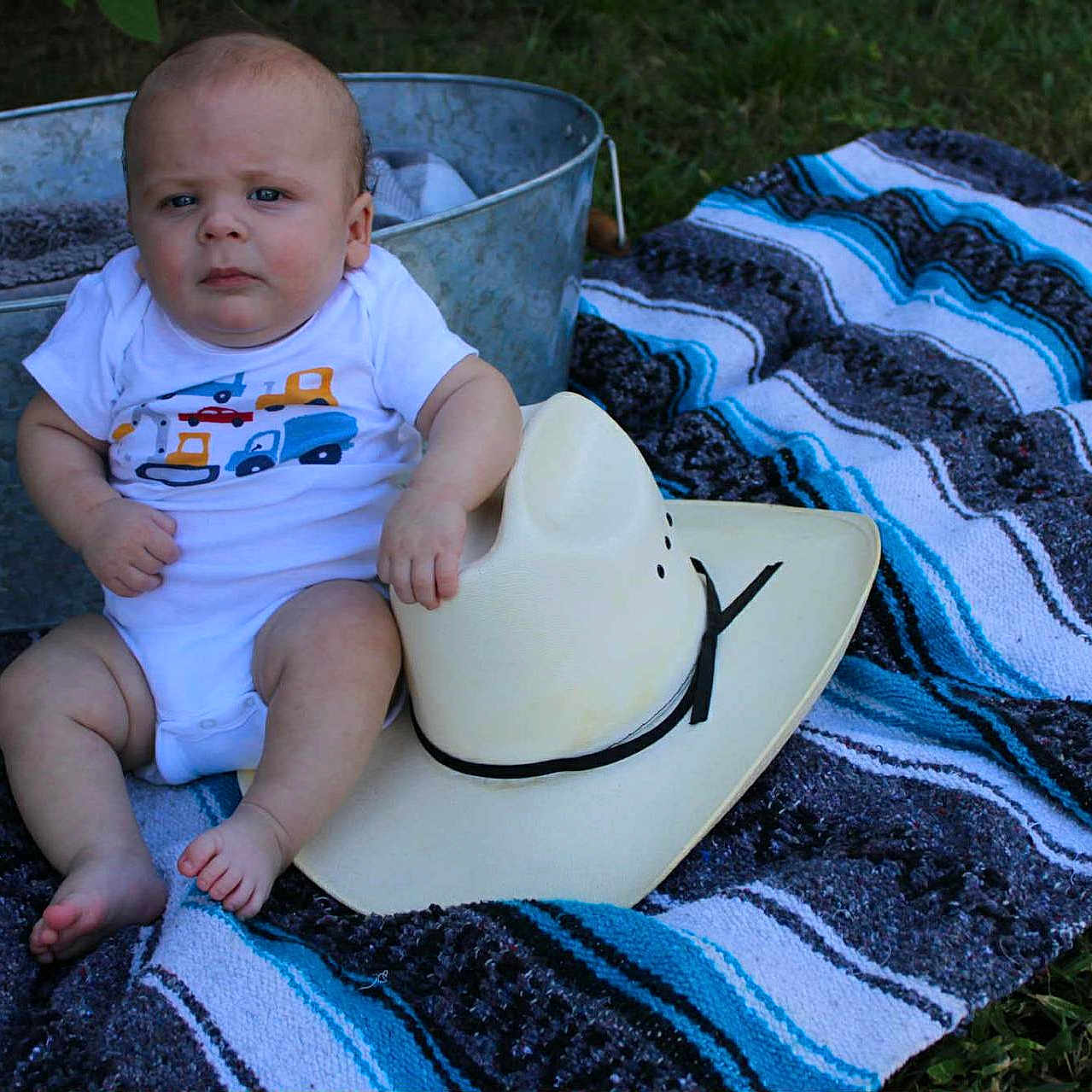 Braxton is registered to the contest to win money with this photo: baby, blanket, blue, clothing, cowboy_hat, cute, foot, grass, gray, hand, infant, metal_tub, outdoor, person, plush_toy, portrait, sitting, striped, summer, toy