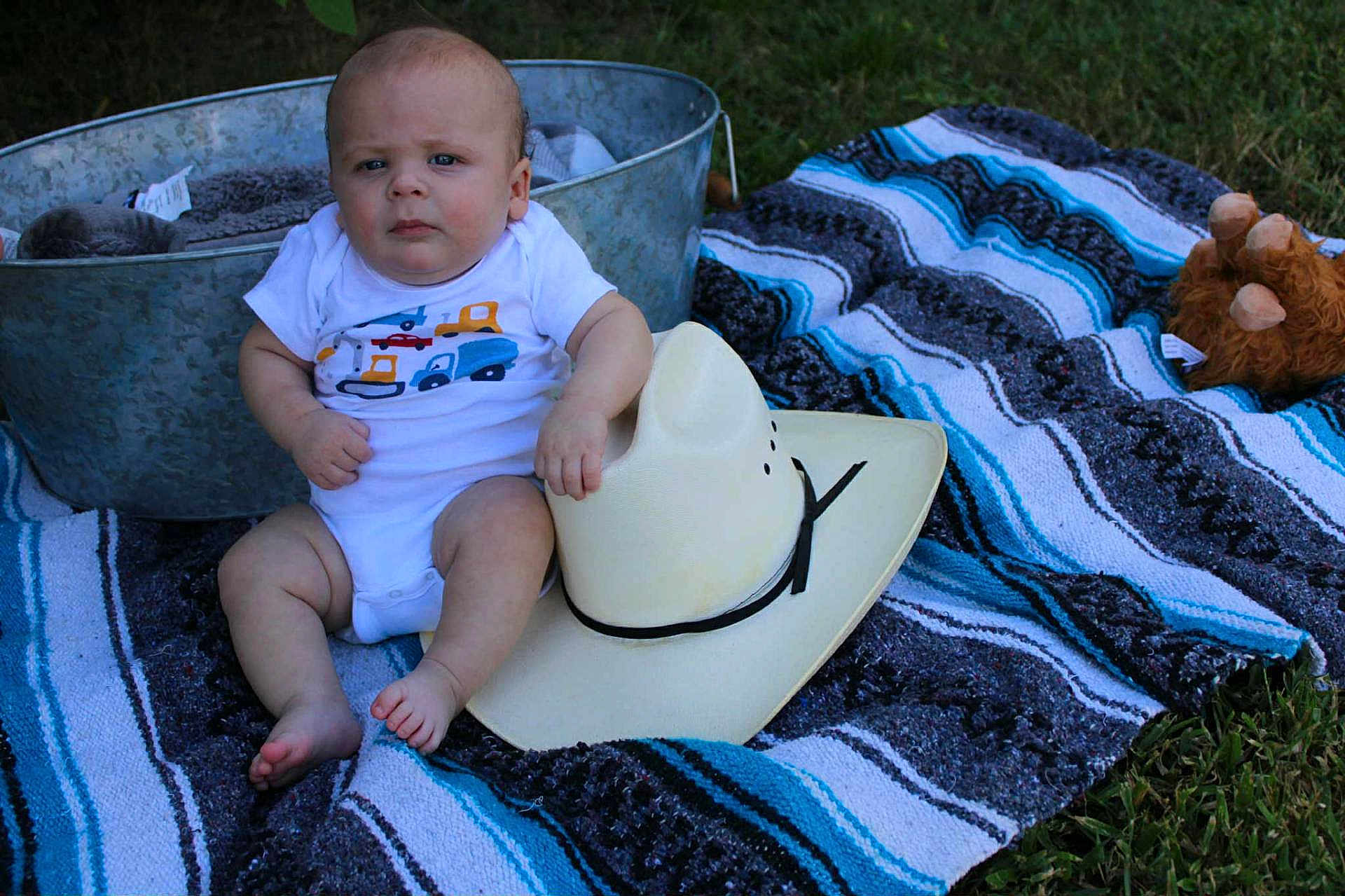 Braxton is registered to the contest to win money with this photo: baby, cowboy_hat, blanket, outdoor, grass, metal_tub, plush_toy, blue, gray, striped, sitting, infant, clothing, foot, hand, person, cute, portrait, summer, toy