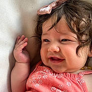 Mia joined the competition — help win amazing prizes! baby, child, smile, curly_hair, bow, pink_bow, coral_dress, floral_pattern, hand, face, cheek, cushion, close_up, happy, infant, skin, portrait, resting, indoors, cute