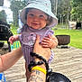 Mia is registered to the contest to win money with this photo: baby, child, hat, purple_dress, tattoo, arm, hand, outdoor, deck, greenery, trees, smile, cute, sun_hat, person, holding, happy, infant, grass, daylight
