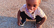 Hailee is registered to the contest to win money with this photo: toddler, child, pacifier, braided_hair, beads, outdoor, gravel, sunlight, sneakers, clothing, curious, crouching, person, shadow, playground, daytime, young_child, cute, expression, casual