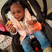 Hailee is registered to the contest to win money with this photo: toddler, child, car_seat, cookie, sippy_cup, pink_vest, jeans, indoor, happy, smiling, person, baby, hair_puffs, clothing, seatbelt, floor, toy, hand, face, casual