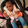 toddler, child, car_seat, cookie, sippy_cup, pink_vest, jeans, indoor, happy, smiling, person, baby, hair_puffs, clothing, seatbelt, floor, toy, hand, face, casual
