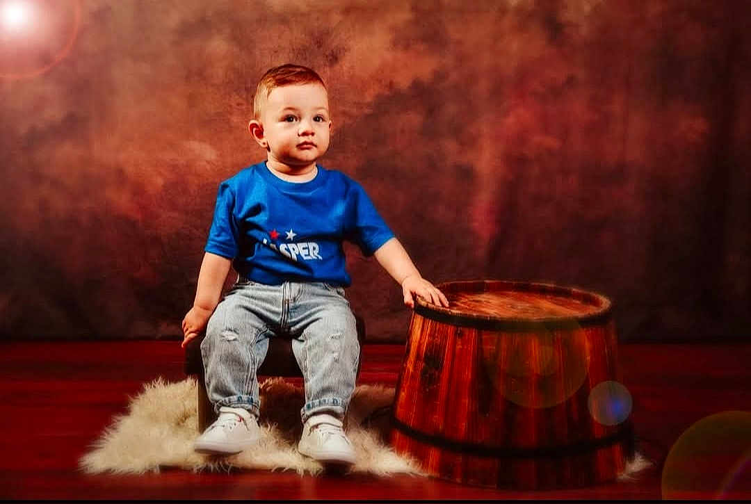 Jasper is registered to the contest to win money with this photo: toddler, child, blue_tshirt, jeans, white_sneakers, wooden_barrel, chair, fluffy_rug, studio, portrait, indoor, serious_expression, brown_background, floor, young_child, casual_clothing, sitting, hand_on_barrel, cute, small_chair