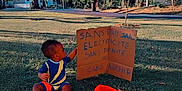 Layhann participe au concours pour gagner de l'argent avec cette photo : toddler, child, grass, outdoor, cardboard, sign, flag, colorful_clothing, blue_sky, trees, park, daylight, sitting, young_child, handwritten_text, nature, sunlight, casual, curious, playful