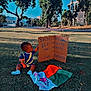 toddler, child, grass, outdoor, cardboard, sign, flag, colorful_clothing, blue_sky, trees, park, daylight, sitting, young_child, handwritten_text, nature, sunlight, casual, curious, playful