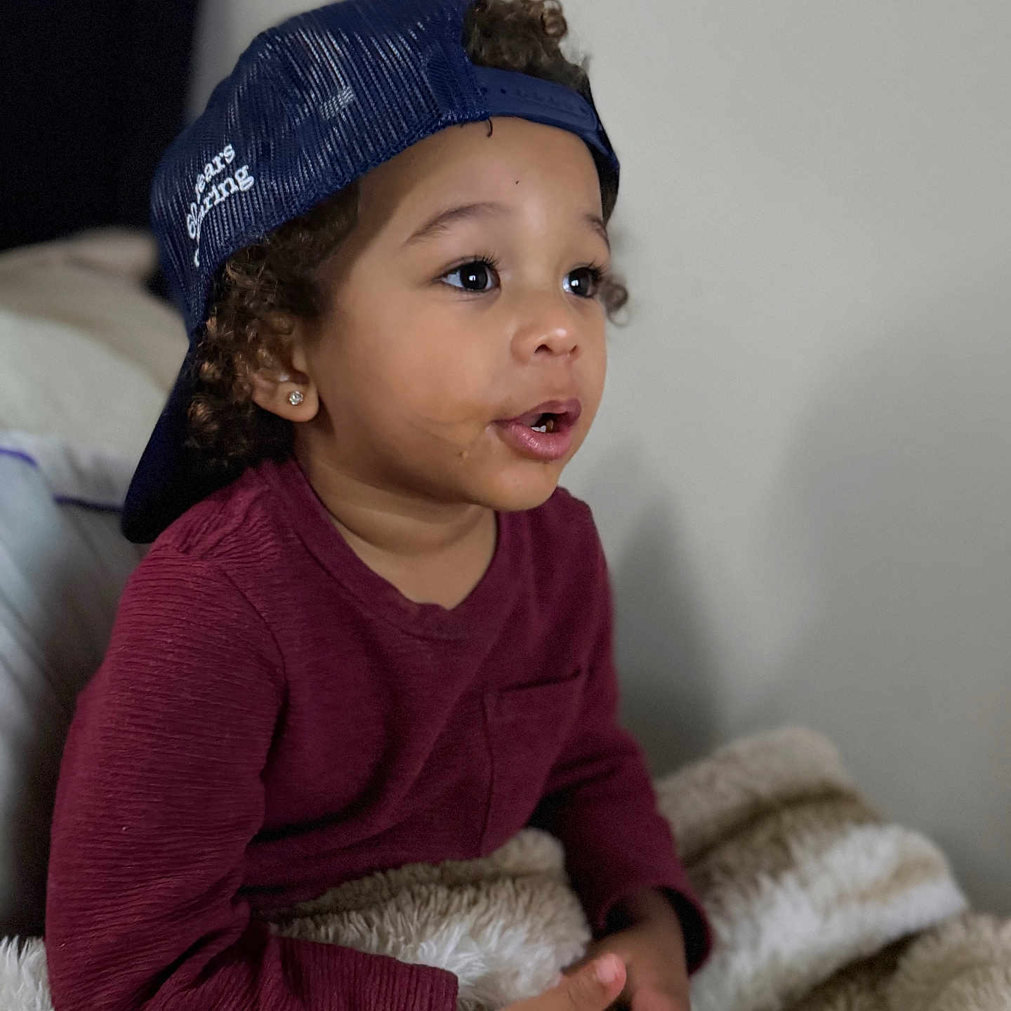 Jakari is registered to the contest to win money with this photo: toddler, child, cap, hat, curly_hair, maroon_shirt, blanket, indoor, portrait, cozy, expression, curious, sitting, young_child, face, earrings, hands, neutral_background, closeup, person