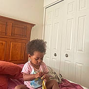 Evelyn is registered to the contest to win money with this photo: toddler, child, bed, blanket, purple, wooden_headboard, closet, door, indoor, person, clothing, pink_shirt, curly_hair, baby, foot, sitting, playing, room, home, furniture