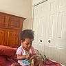 toddler, child, bed, blanket, purple, wooden_headboard, closet, door, indoor, person, clothing, pink_shirt, curly_hair, baby, foot, sitting, playing, room, home, furniture