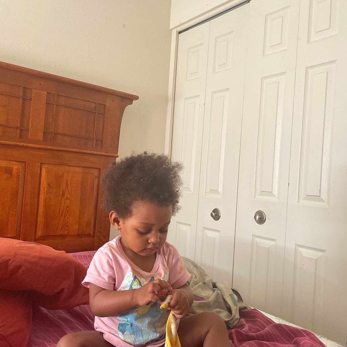 Evelyn is registered to the contest to win money with this photo: toddler, child, bed, blanket, purple, wooden_headboard, closet, door, indoor, person, clothing, pink_shirt, curly_hair, baby, foot, sitting, playing, room, home, furniture