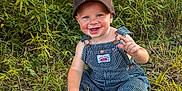 Karter joined the competition — help win amazing prizes! toddler, child, smiling, grass, greenery, outdoor, cap, overalls, happy, nature, baby, sitting, footwear, plants, sunlight, face, portrait, cute, young_child, clothing