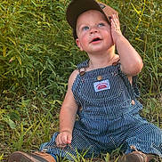 Karter is registered to the contest to win money with this photo: toddler, child, grass, outdoor, nature, greenery, overalls, cap, blue_eyes, happy, sitting, curious, expression, casual_clothing, footwear, plants, daylight, portrait, person, playful
