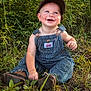 Karter is registered to the contest to win money with this photo: toddler, child, smiling, overalls, cap, outdoor, grass, greenery, happy, baby, nature, sitting, cute, portrait, young_child, daylight, casual_clothing, cheerful, person, headwear