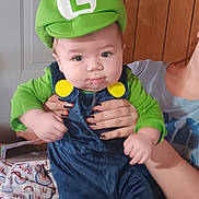 Easton is registered to the contest to win money with this photo: baby, child, costume, luigi_hat, green_hat, blue_overalls, yellow_buttons, drool, person_holding, ripped_jeans, wooden_wall, door, indoor, curious_expression, hands, sitting, clothing, portrait, face, cute