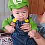 baby, blue_overalls, child, clothing, costume, curious_expression, cute, door, drool, face, green_hat, hands, indoor, luigi_hat, person_holding, portrait, ripped_jeans, sitting, wooden_wall, yellow_buttons