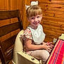 Izabella joined the competition — help win amazing prizes! child, smiling, bow, bib, booster_seat, table, tablecloth, wooden_wall, wooden_chair, indoor, happy, person, face, furniture, clothing, pink_nail_polish, sitting, cute, young_child, cozy