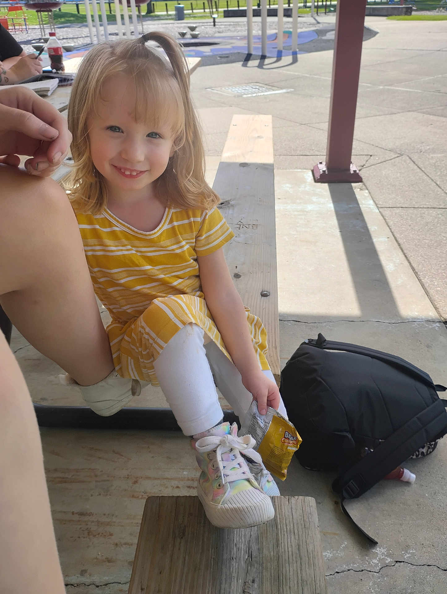 Izabella is registered to the contest to win money with this photo: child, girl, smile, yellow_dress, white_leggings, sneakers, snack, bench, outdoor, playground, backpack, sunlight, person, happy, wood, park, casual, holding, sitting, daytime