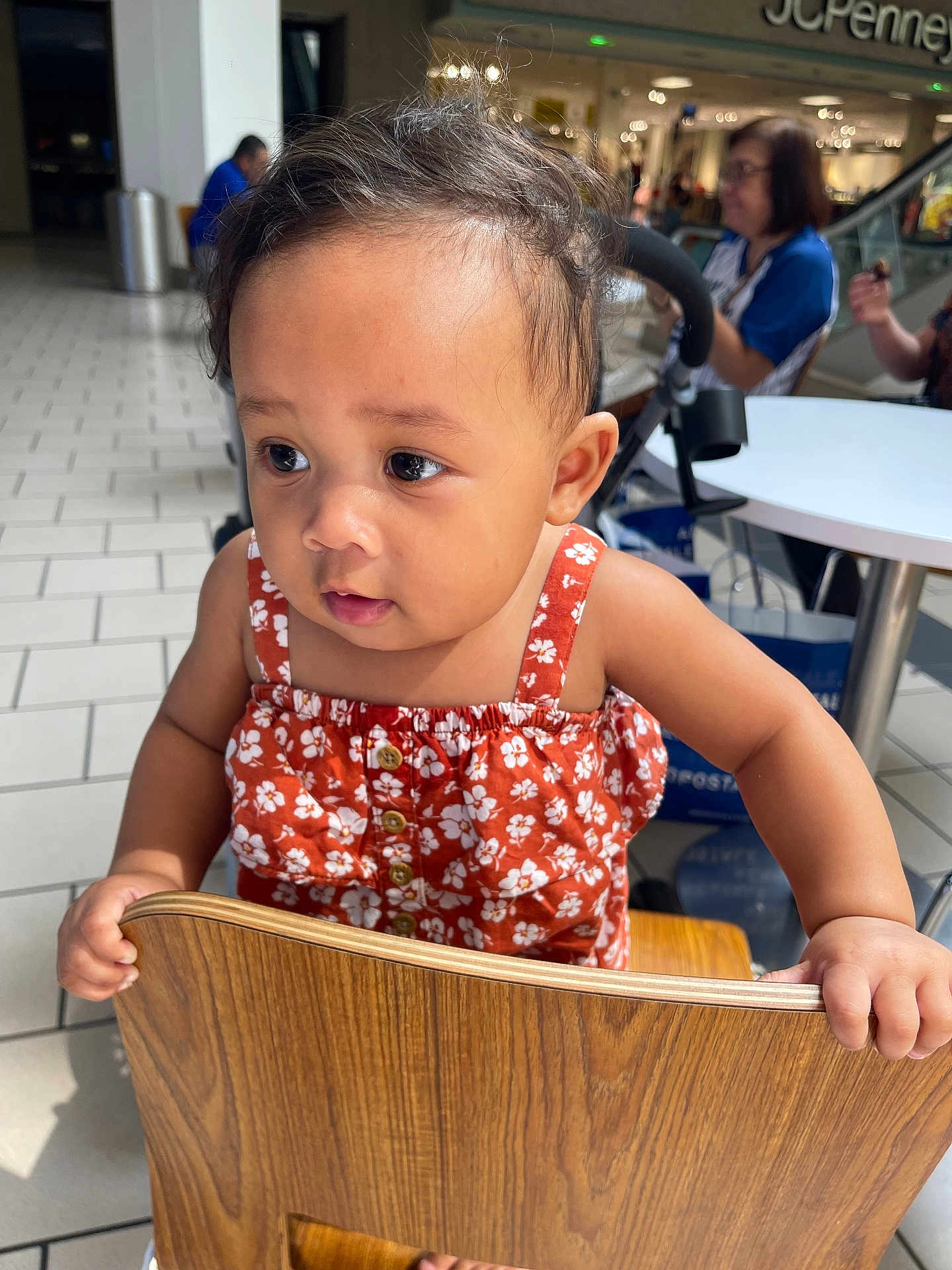 Amelia is registered to the contest to win money with this photo: toddler, child, orange_dress, floral_pattern, wooden_chair, shopping_mall, food_court, table, people, background, curious, indoor, floor_tiles, person, holding, small_hands, cute, casual_wear, daylight, portrait
