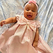 Amelia is registered to the contest to win money with this photo: baby, smiling, pink_dress, headband, bow, lying_down, floral_sheet, happy, infant, child, cute, portrait, indoors, fabric, pattern, person, celebration, toddler, joyful, skin