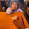Nova is registered to the contest to win money with this photo: casual, child, clothing, cup, curly_hair, cute, eating, food, hair, hand, happy, high_chair, indoor, messy_face, person, restaurant, sleeves, table, toddler, wooden_table