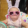 baby, child, sunglasses, pink, bunny_ears, walker, toy, indoor, smile, cute, chubby_cheeks, colorful, person, headwear, infant, clothing, baby_toy, face, seat, comfort