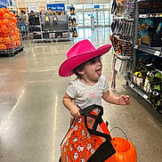 Maeve joined the competition — help win amazing prizes! toddler, child, pink_hat, cowboy_hat, halloween, pumpkin_bucket, shopping_bag, store_interior, aisle, toys, decorations, floor, smiling, playful, curly_hair, clothing, retail, holiday, fun, person