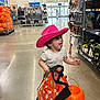 toddler, child, pink_hat, cowboy_hat, halloween, pumpkin_bucket, shopping_bag, store_interior, aisle, toys, decorations, floor, smiling, playful, curly_hair, clothing, retail, holiday, fun, person
