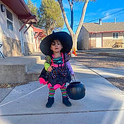 Maeve is registered to the contest to win money with this photo: child, costume, witch_hat, pumpkin_bucket, sidewalk, outdoor, daylight, tree, building, striped_tights, black_shoes, colorful_dress, curly_hair, holiday, halloween, playful, person, young_child, residential_area, sunny