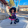 child, costume, witch_hat, pumpkin_bucket, sidewalk, outdoor, daylight, tree, building, striped_tights, black_shoes, colorful_dress, curly_hair, holiday, halloween, playful, person, young_child, residential_area, sunny