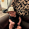 baby, child, infant, sitting, chair, leather_chair, baseball_cap, sneakers, black_clothing, leopard_print, cushion, lamp, indoor, portrait, fashion, cute, smiling, face, furniture, home_decor