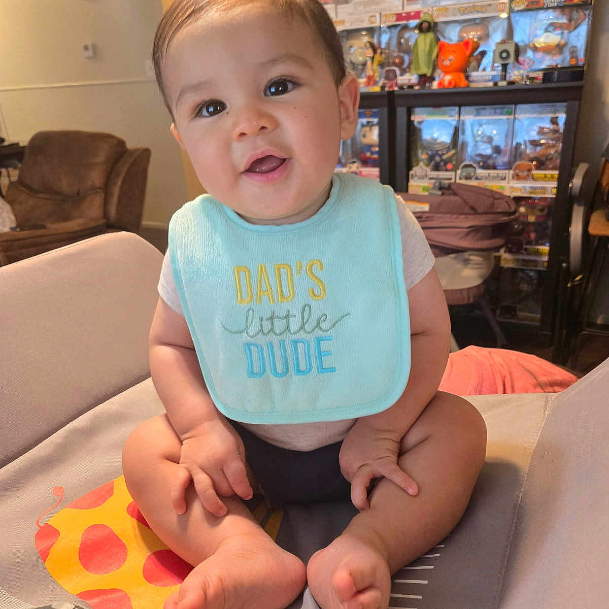 Jino is registered to the contest to win money with this photo: baby, baby_seat, bib, child, clothing, collectibles, cute, face, feet, funko_pops, furniture, giraffe, happy, indoors, infant, living_room, person, sitting, smiling, toys