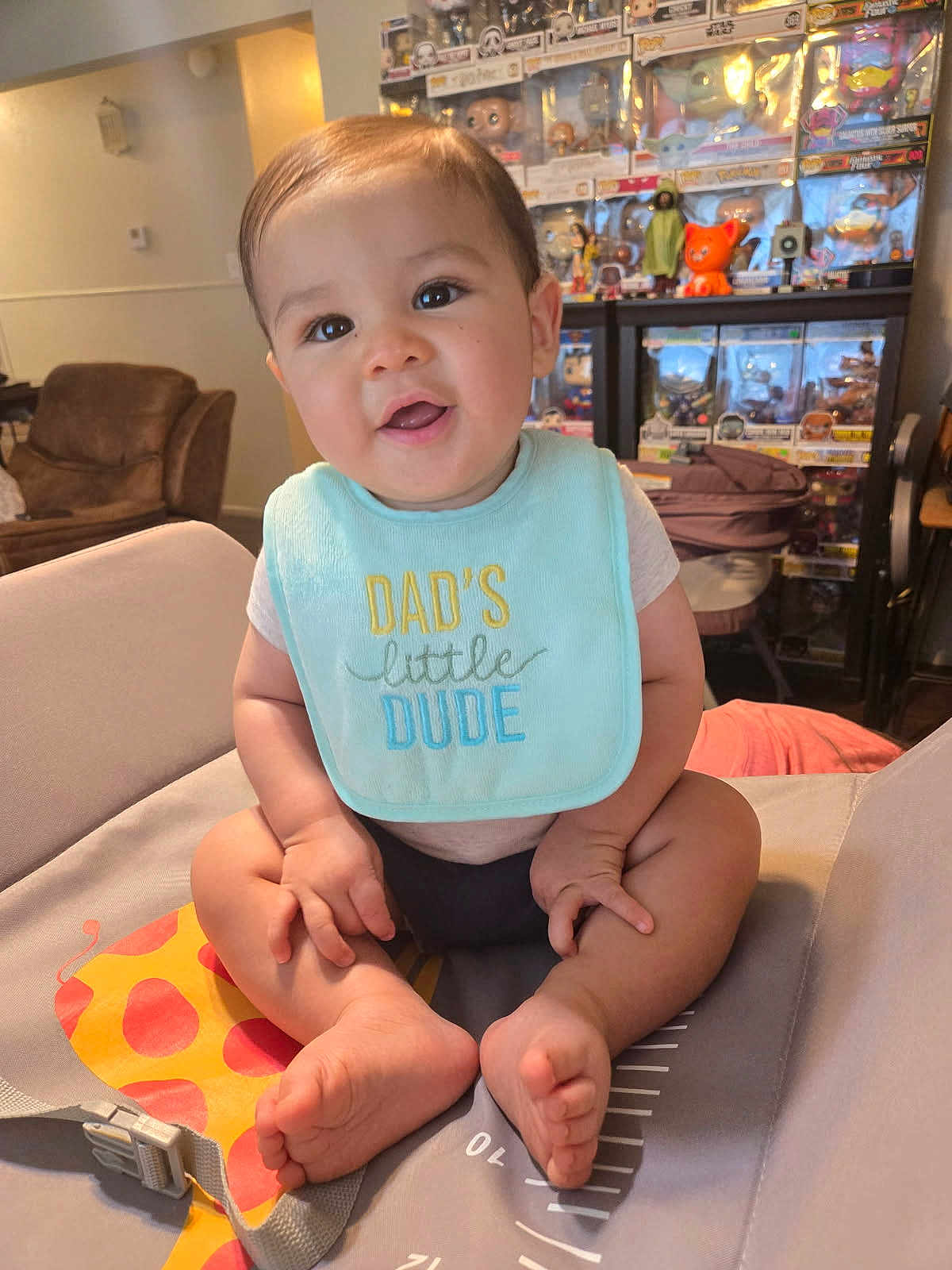 Jino is registered to the contest to win money with this photo: baby, child, infant, smiling, bib, sitting, giraffe, baby_seat, living_room, toys, collectibles, funko_pops, furniture, feet, cute, happy, indoors, person, clothing, face