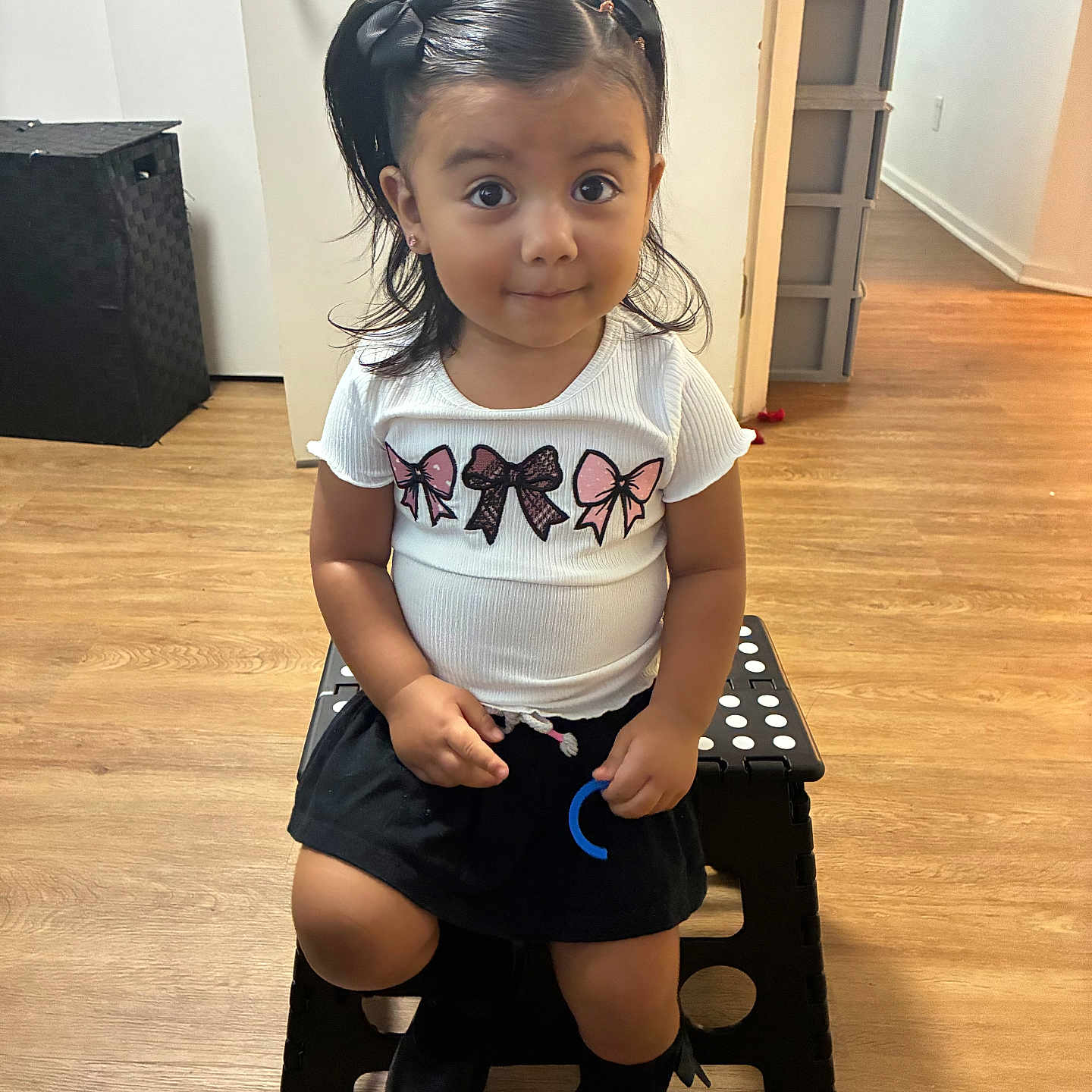 Maricela is registered to the contest to win money with this photo: child, girl, pigtails, black_bows, white_shirt, black_skirt, boots, toy, step_stool, wooden_floor, indoor, door, wall, curious_expression, sitting, person, young_child, cute, smile, hair
