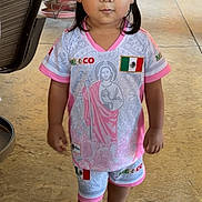 Maricela is registered to the contest to win money with this photo: toddler, child, girl, outdoor, pigtails, bows, sandals, shirt, shorts, mexico_flag, curious_expression, concrete_floor, chair, table, plants, clothing, person, portrait, summer, daylight