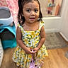 child, girl, dress, yellow, hair_bows, purse, indoor, floor, wooden_floor, smile, face, person, young_child, curly_hair, cute, fashion, portrait, standing, home, happy